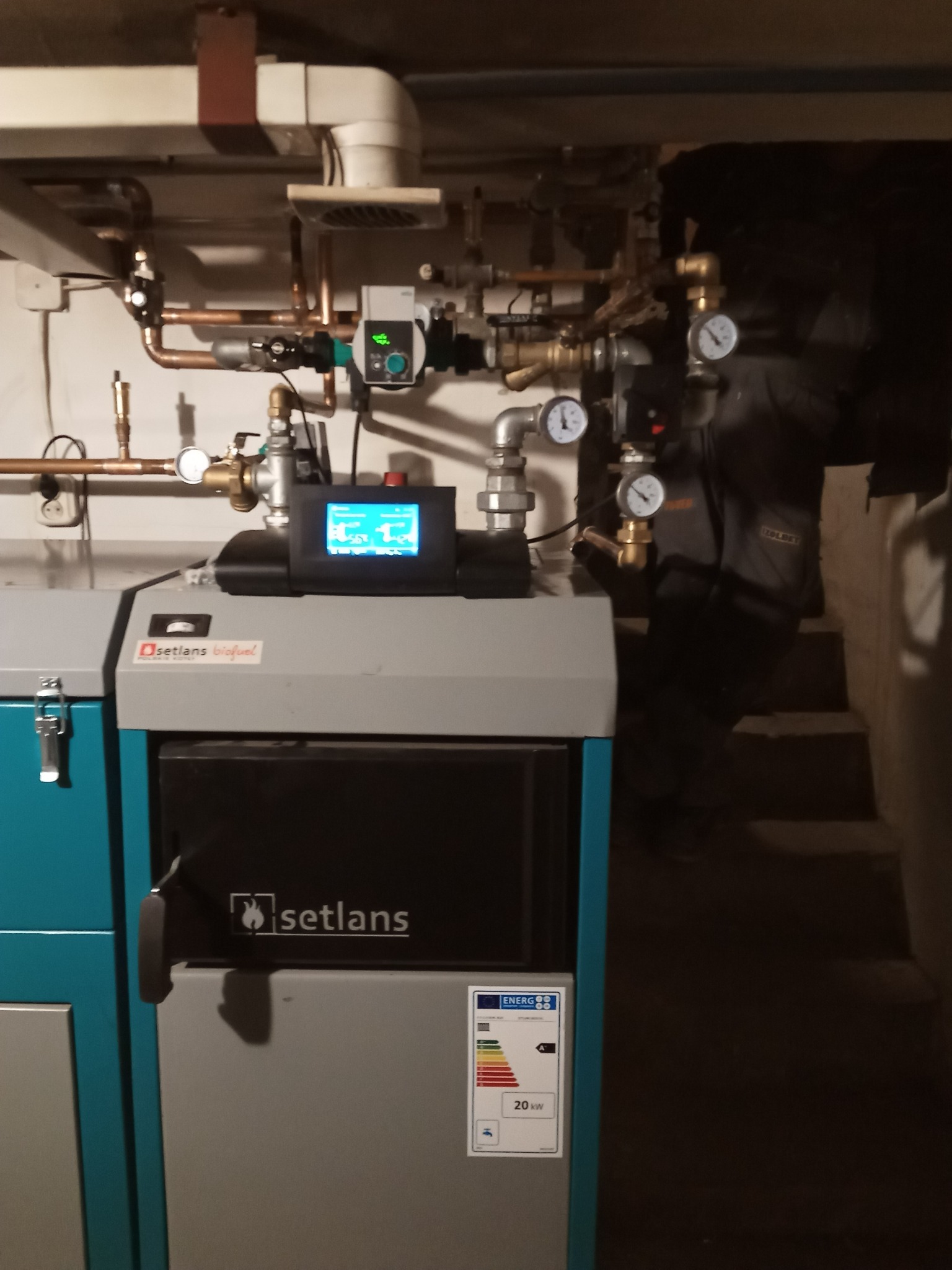 Nowoczesny piec Setlans Biofuel z panelem sterowania i licznymi zaworami oraz manometrami na miedzianych rurach, widoczny w tle pracownik w kombinezonie roboczym.