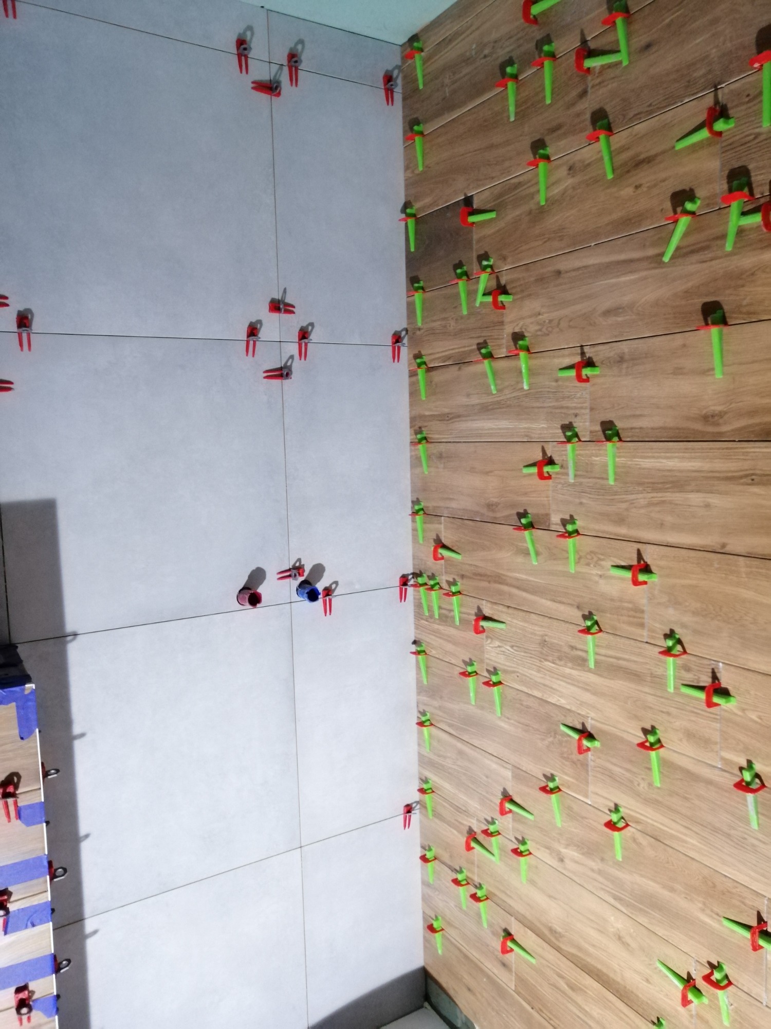 Świeżo ułożone płytki ścienne w dwóch wzorach, jasnoszarym i imitującym drewno, z użyciem systemu poziomowania płytek (zielone kliny i czerwone klipsy), widoczne rurki instalacyjne wystające ze...