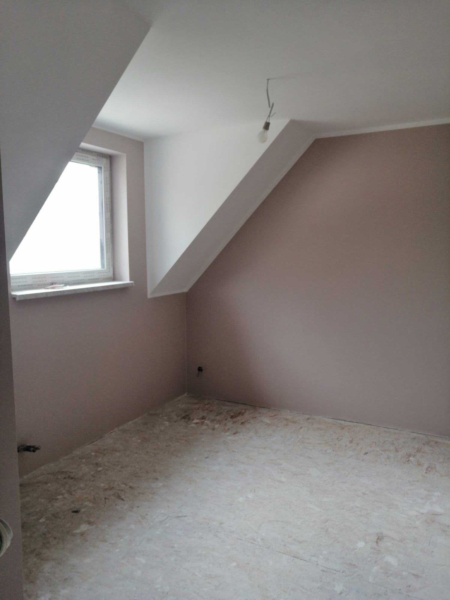 Pokój na poddaszu po malowaniu ścian na kolor pudrowy róż, widoczna surowa podłoga i wystająca żarówka na kablu.