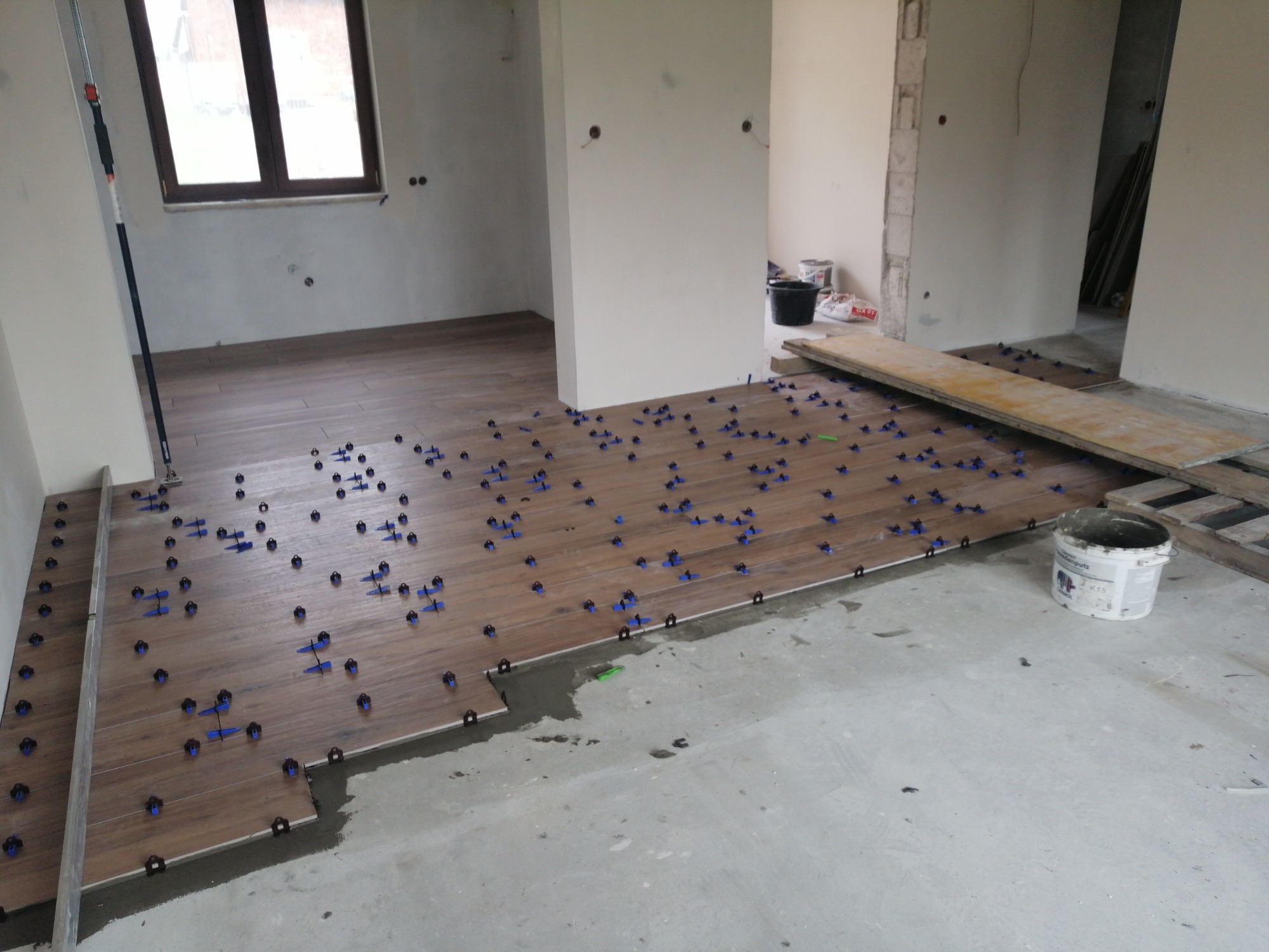 Układanie płytek podłogowych imitujących drewno, z użyciem niebieskich krzyżyków dystansowych, widoczne niedokończone fragmenty i wiadro po kleju.
