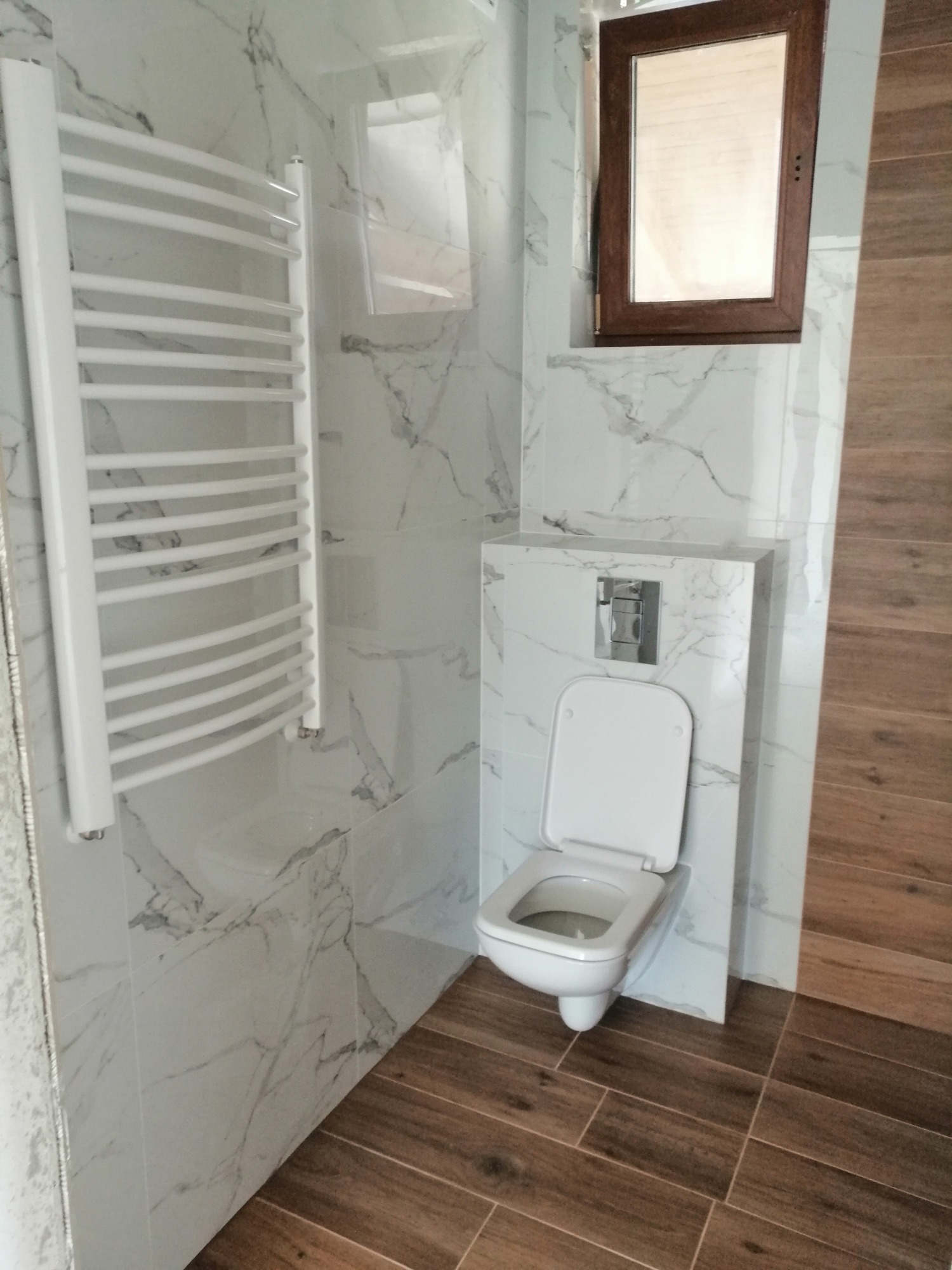 Nowoczesna łazienka z białymi płytkami imitującymi marmur na ścianach, drewnopodobną podłogą, wiszącą toaletą i białym grzejnikiem drabinkowym, oświetlona światłem dziennym wpadającym przez małe okno.