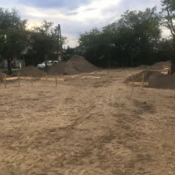 Przygotowany teren pod budowę z wyznaczonymi punktami geodezyjnymi, widoczne pryzmy ziemi i drzewa w tle.