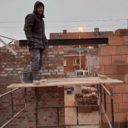 Mężczyzna w kurtce i kapturze stoi na rusztowaniu, murowana ściana z czerwonej cegły w trakcie budowy, widoczna stalowa belka nad otworem okiennym, w tle zabudowa mieszkalna.