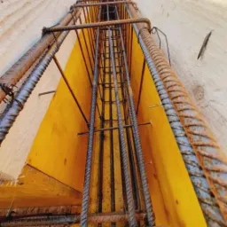 Zbrojenie pionowe w szalunku fundamentowym z żółtą sklejką, widok z dołu w górę, z widocznymi prętami żebrowanymi i elementami mocującymi.