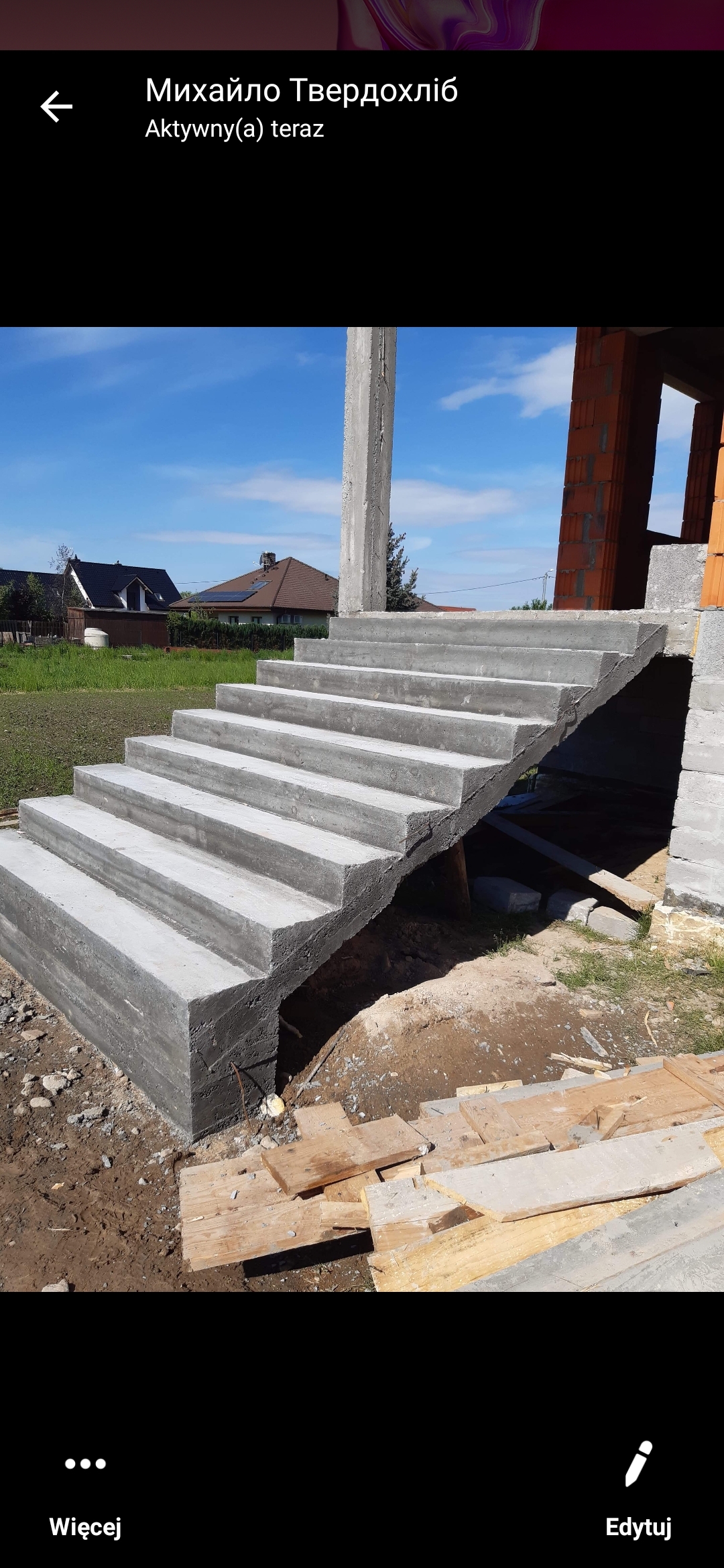 Surowe betonowe schody zewnętrzne prowadzące do wznoszonego domu z czerwonej cegły, widoczne elementy szalunku, w tle trawnik i domy jednorodzinne.