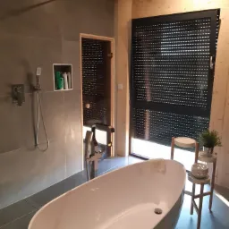 Nowoczesna łazienka z wolnostojącą wanną, szarą armaturą i drewnianymi elementami wykończeniowymi, rolety zewnętrzne na oknie.