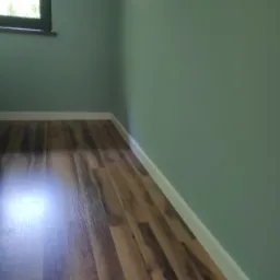 Świeżo pomalowane, zielone ściany w pokoju z ułożonymi panelami podłogowymi o wzorze drewna i białymi listwami przypodłogowymi, widoczne okno z ciemną ramą.