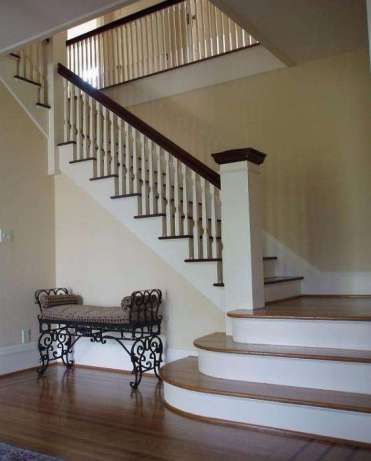 Jasne wnętrze z drewnianymi schodami z białymi balustradami i ciemnobrązowymi poręczami, dekoracyjna ławka z kutego żelaza pod schodami.