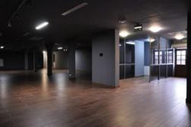 Pusta, ciemna przestrzeń biurowa z drewnianą podłogą, szarymi ścianami i kolumnami. Oświetlenie jarzeniowe na suficie i przeszklone boksy biurowe w tle.