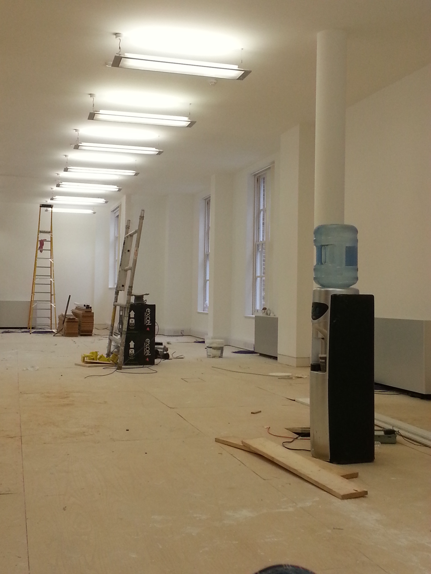 Przygotowania do malowania w jasnym biurze: widoczne drabiny, puszki z farbą i dystrybutor wody na tle białych ścian i podłogi pokrytej płytami OSB.