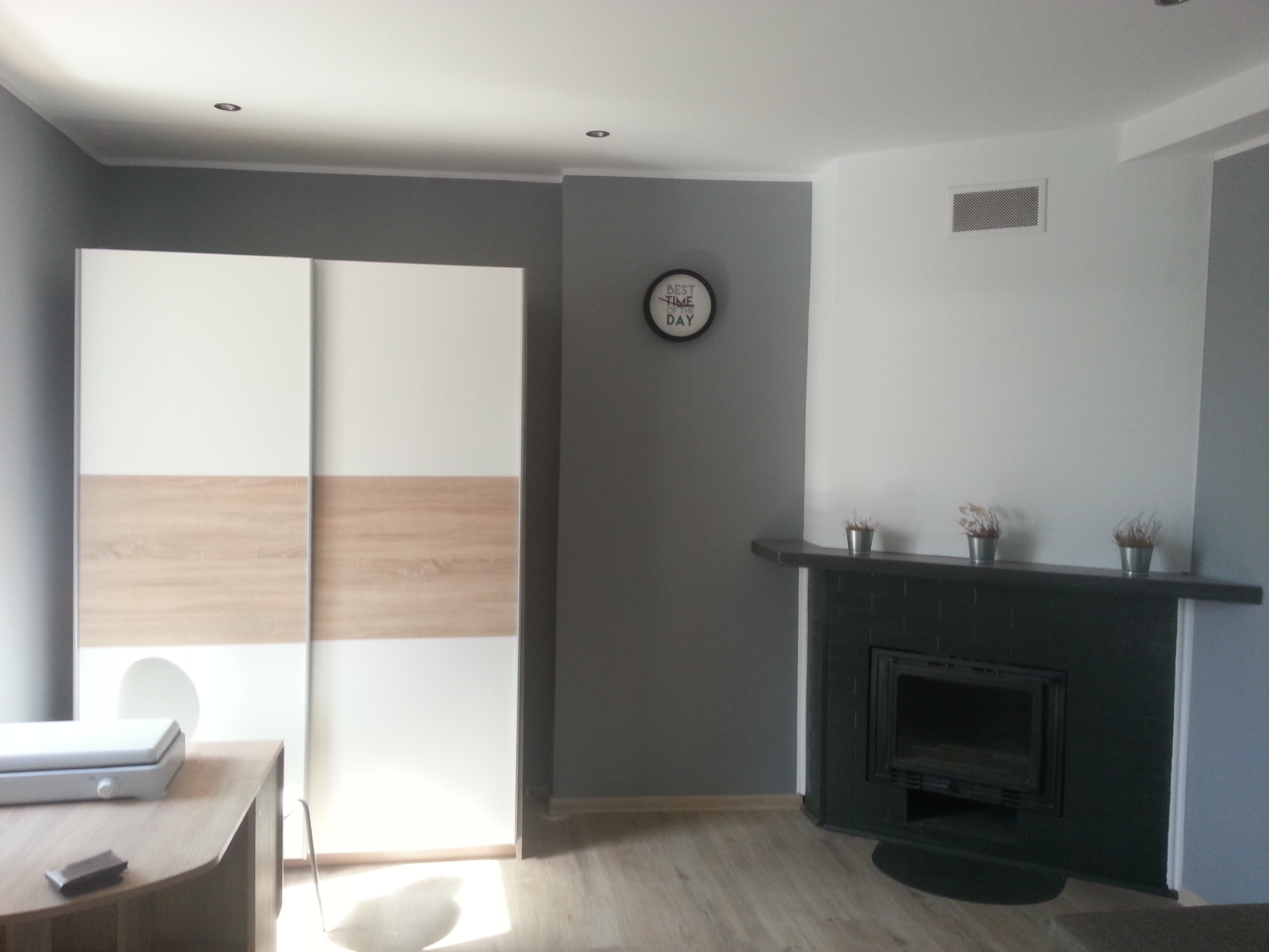 Pokój po remoncie: ściany pomalowane na szaro i biało, biało-drewniana szafa przesuwna, czarny kominek z trzema doniczkami na górze, zegar na ścianie.