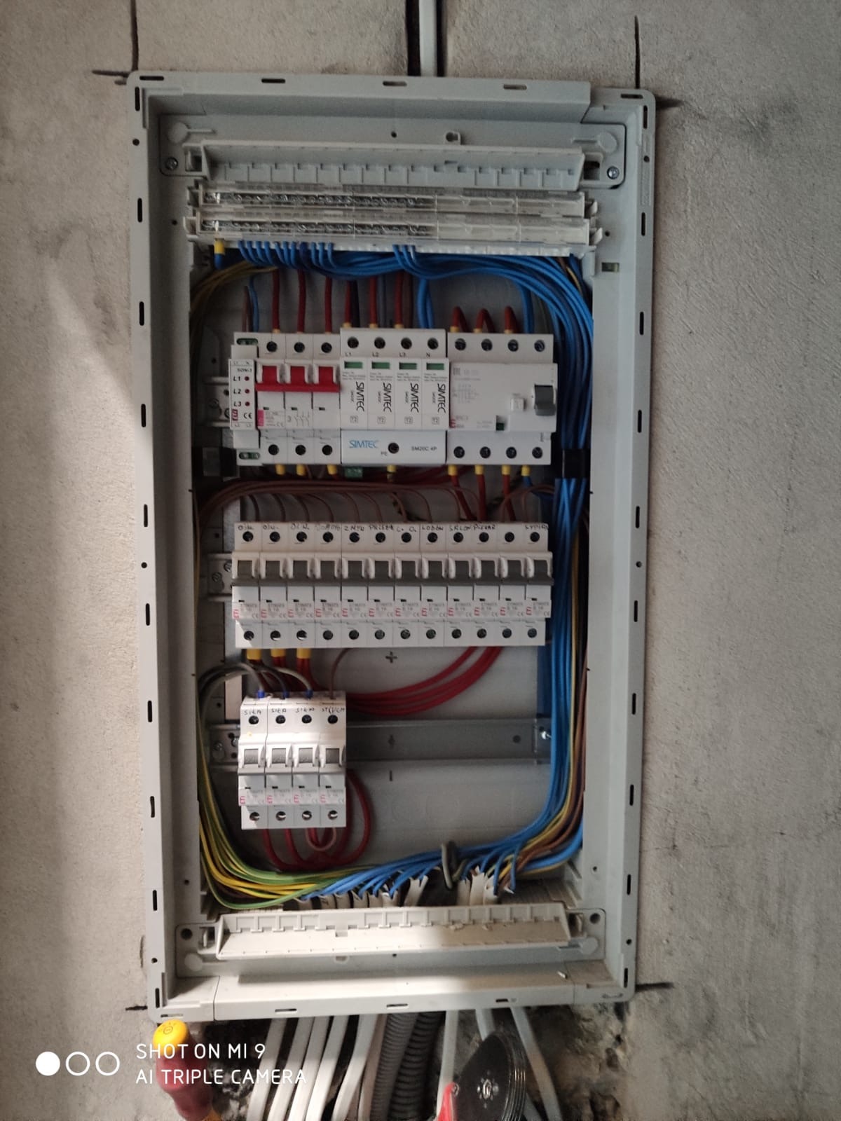 Otwarta skrzynka rozdzielcza z zainstalowanymi bezpiecznikami i przewodami w kolorach niebieskim, czerwonym i żółto-zielonym, zamontowana na surowej, szarej ścianie.