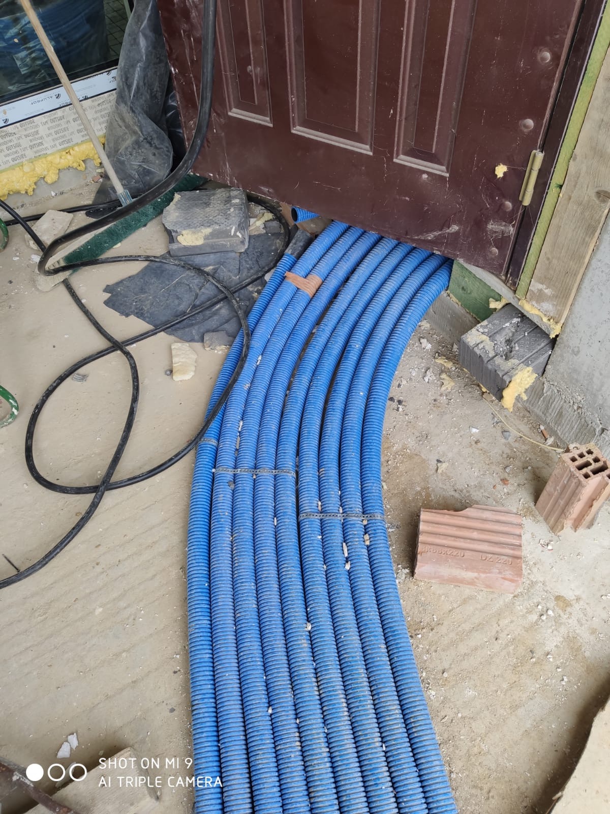 Kłębek niebieskich rur karbowanych wchodzących pod próg drzwi, widoczne kable elektryczne i gruz budowlany.