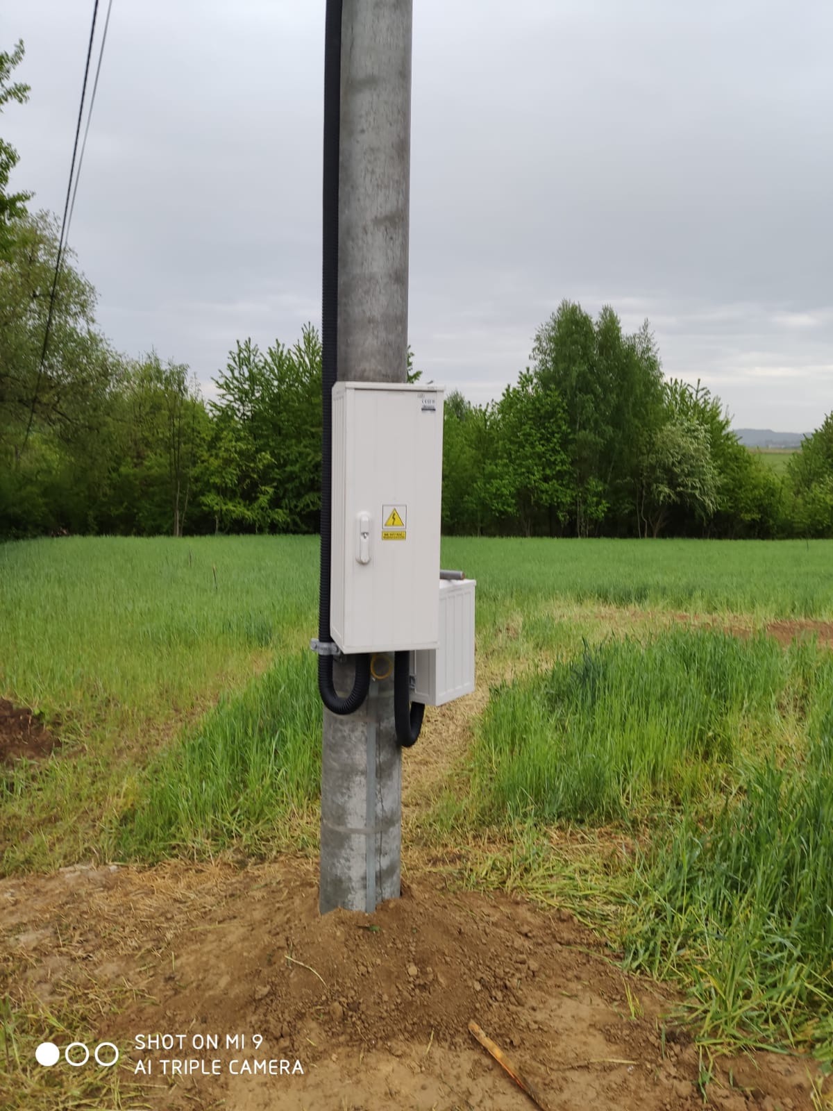 Słup energetyczny z zamontowaną białą skrzynką elektryczną z symbolem ostrzegawczym, widoczne czarne rury osłonowe, w tle łąka i drzewa pod pochmurnym niebem.