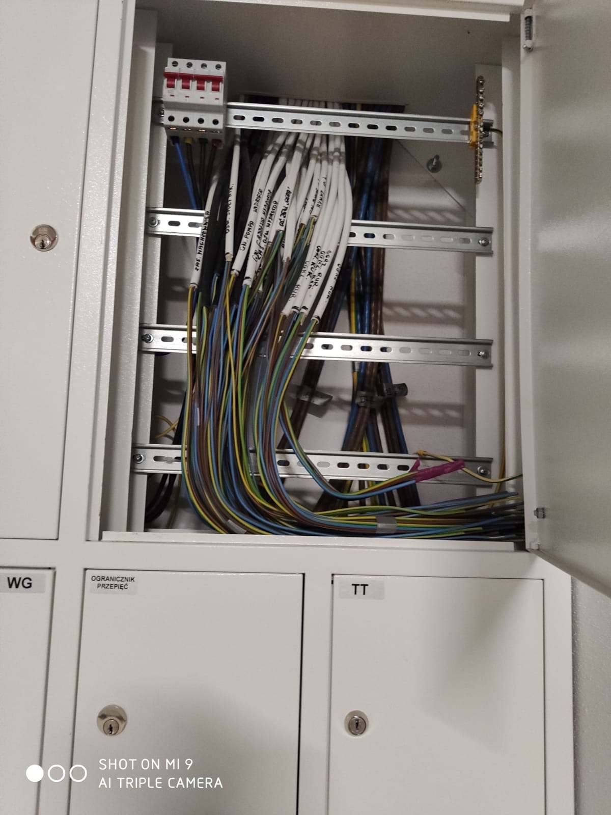 Otwarta skrzynka elektryczna z licznymi przewodami oznaczonymi etykietami, ułożonymi na metalowych szynach, widoczne zabezpieczenia nad przewodami.