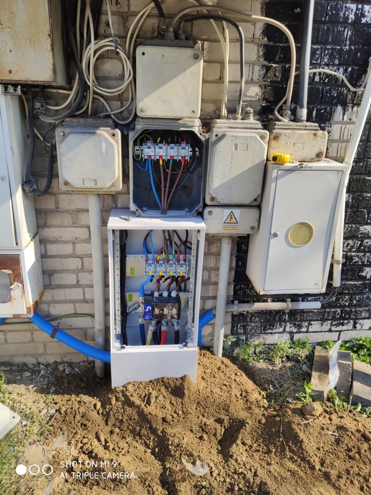 Otwarta skrzynka elektryczna z widocznymi przewodami i bezpiecznikami, zamontowana na ceglanej ścianie obok innych, starszych skrzynek. Wokół ziemia i fragment niebieskiej rury karbowanej.