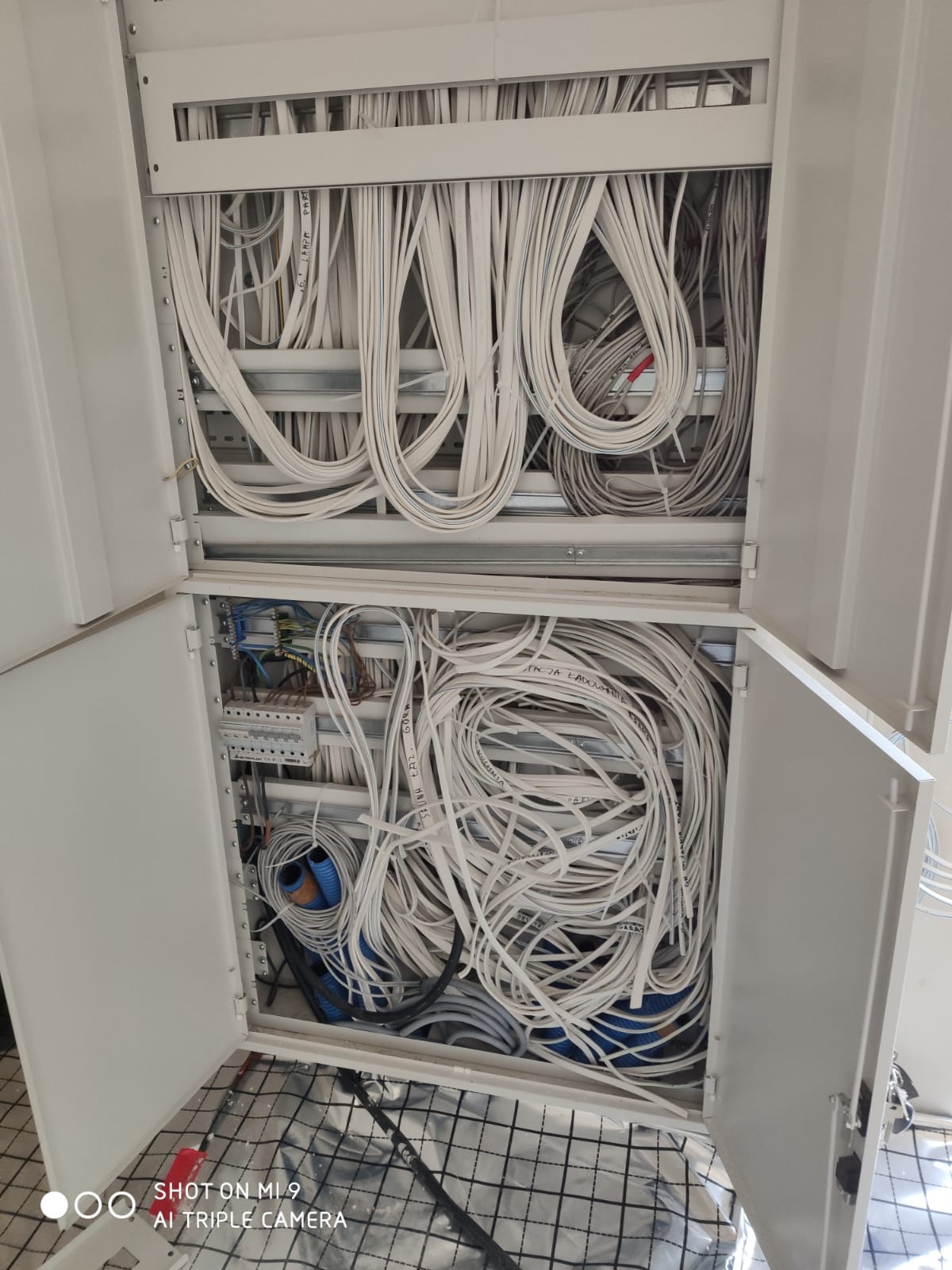 Otwarta szafa rozdzielcza z chaotycznie ułożonymi przewodami elektrycznymi, widoczne złącza i listwy zaciskowe, przewody w kolorze białym i niebieskim, dolna część szafy na tle podłogi wyłożonej...