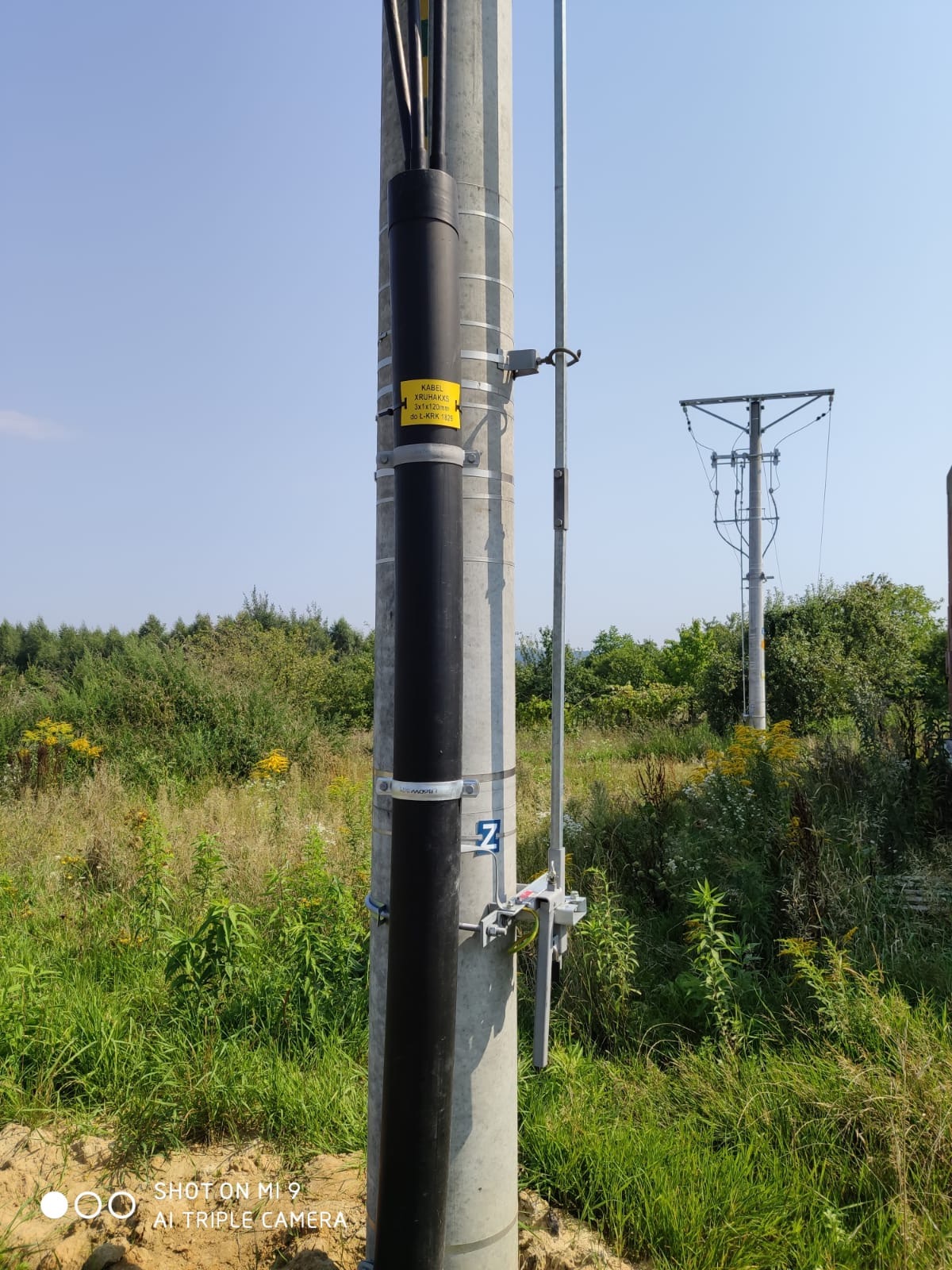 Słup energetyczny z zamontowaną rurą osłonową na kable, zabezpieczoną metalowymi obejmami i żółtą naklejką informacyjną, na tle łąki i błękitnego nieba.