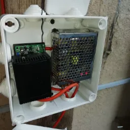 Otwarta biała puszka elektryczna z zainstalowanymi urządzeniami: zasilacz impulsowy w metalowej obudowie z perforacją, płytka drukowana z elementami elektronicznymi i czarny moduł, połączone...