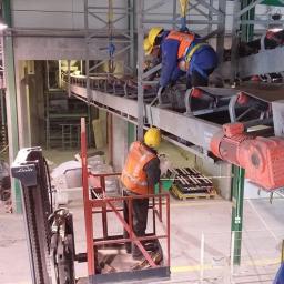 Br Emst Group Sp. z o. o. - Dwóch robotników w kaskach i kamizelkach ochronnych pracuje na wysokości przy taśmociągu przemysłowym, jeden na podnośniku Linde, drugi zabezpieczony uprzężą, widoczny silnik elektryczny...