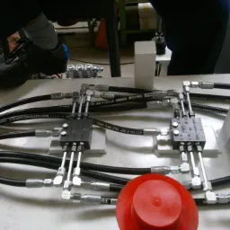 Hydrauliczny system z przewodami, złączkami i zaworami na białej powierzchni, z widoczną stopą w czarnym bucie i szarej skarpetce w tle.