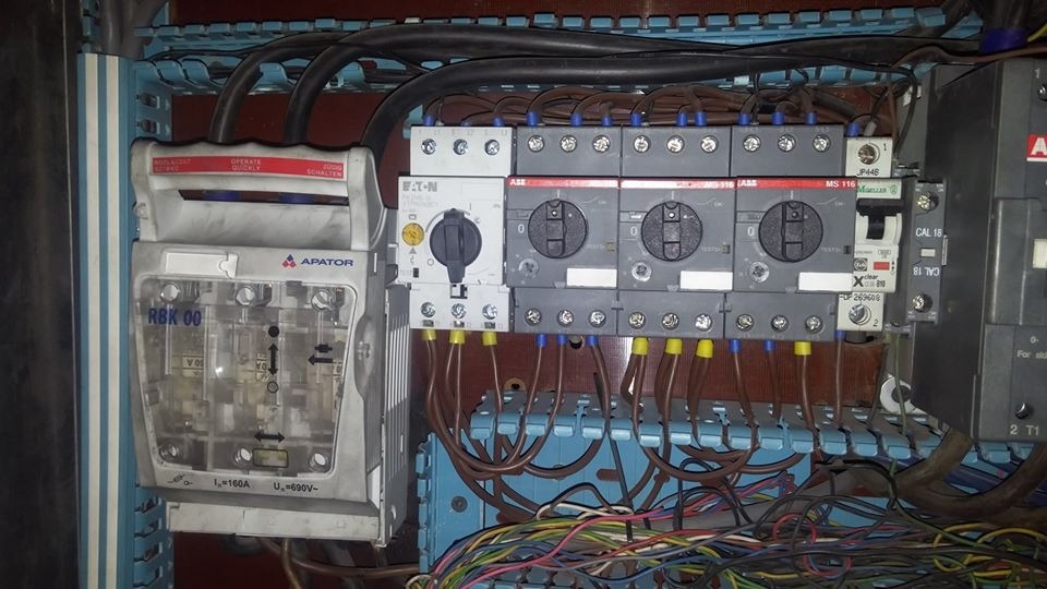 Rozdzielnica elektryczna z widocznymi bezpiecznikami, wyłącznikami i przewodami w kolorach żółtym, niebieskim i brązowym, marki Apator i Eaton.