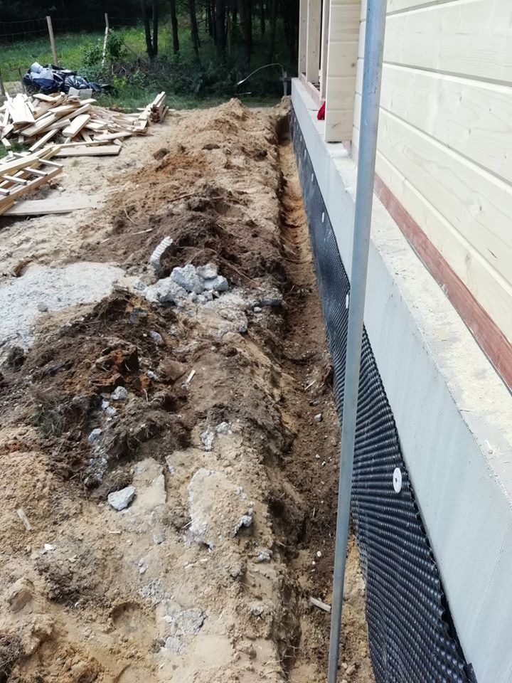 Wykop w ziemi wzdłuż fundamentu drewnianego domu, zabezpieczonego folią kubełkową, widoczne resztki betonu i deski.