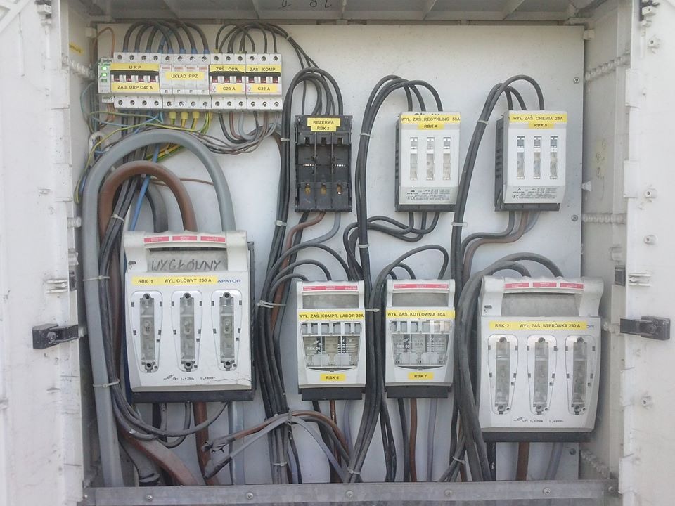 Otwarta skrzynka elektryczna z bezpiecznikami i przewodami, widoczne oznaczenia 'Wyłącznik główny 250A', 'Rezerwa', 'Recykling', 'Chemia', 'Sterówka',  różne zabezpieczenia i układy.