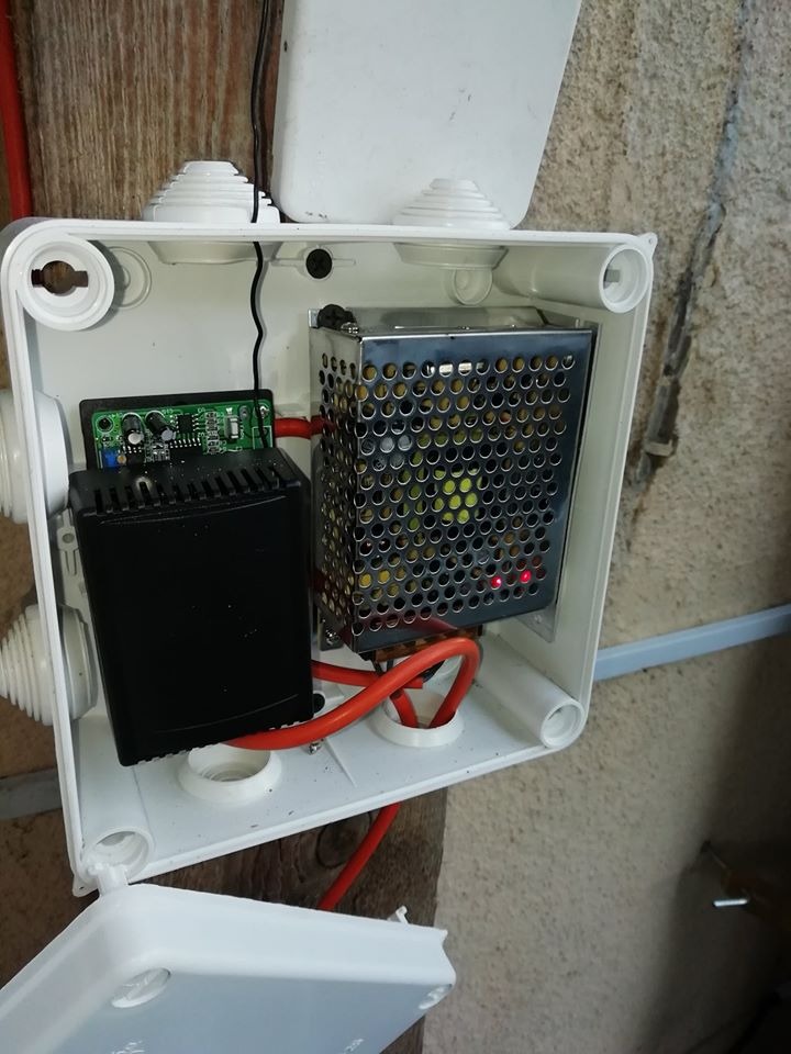 Otwarta biała puszka elektryczna z zainstalowanymi urządzeniami: zasilacz impulsowy w metalowej obudowie z perforacją, płytka drukowana z elementami elektronicznymi i czarny moduł, połączone...