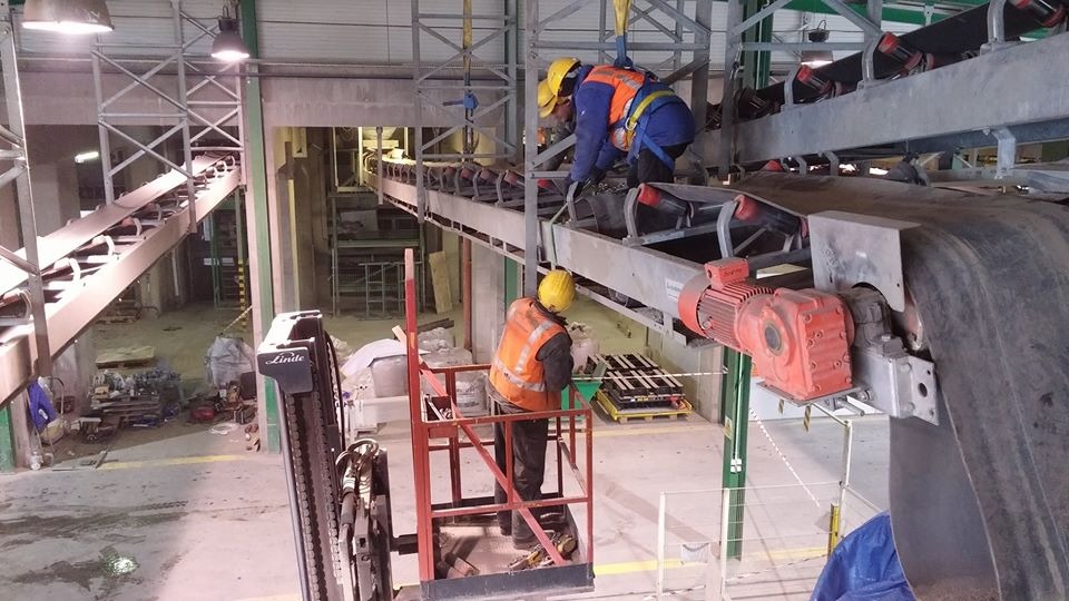 Dwóch robotników w kaskach i kamizelkach ochronnych pracuje na wysokości przy taśmociągu przemysłowym, jeden na podnośniku Linde, drugi zabezpieczony uprzężą, widoczny silnik elektryczny...