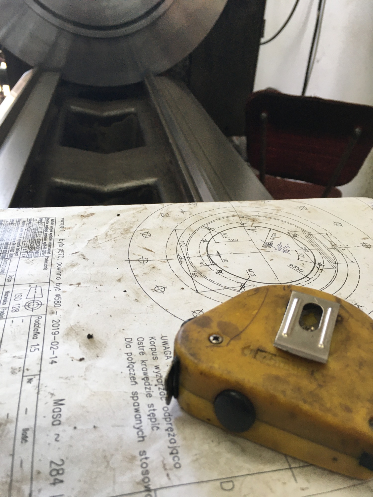 Żółta, zabrudzona miara zwijana leży na technicznym rysunku konstrukcyjnym z uwagami dotyczącymi spawania. W tle widoczny fragment metalowej konstrukcji i czerwone krzesło.