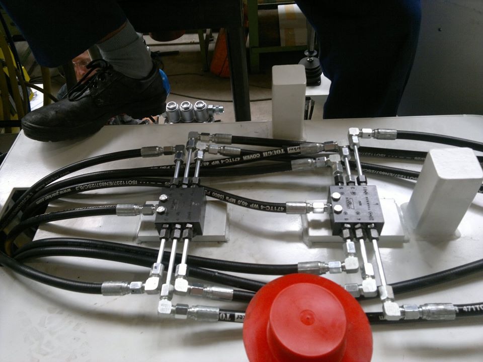 Hydrauliczny system z przewodami, złączkami i zaworami na białej powierzchni, z widoczną stopą w czarnym bucie i szarej skarpetce w tle.