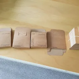 Brązowe, kartonowe opakowania na żarówki LED z nadrukowanym logo i napisem 'Rozświetlamy Twój Dom', ułożone obok stosu pustych, złożonych opakowań związanych gumką.