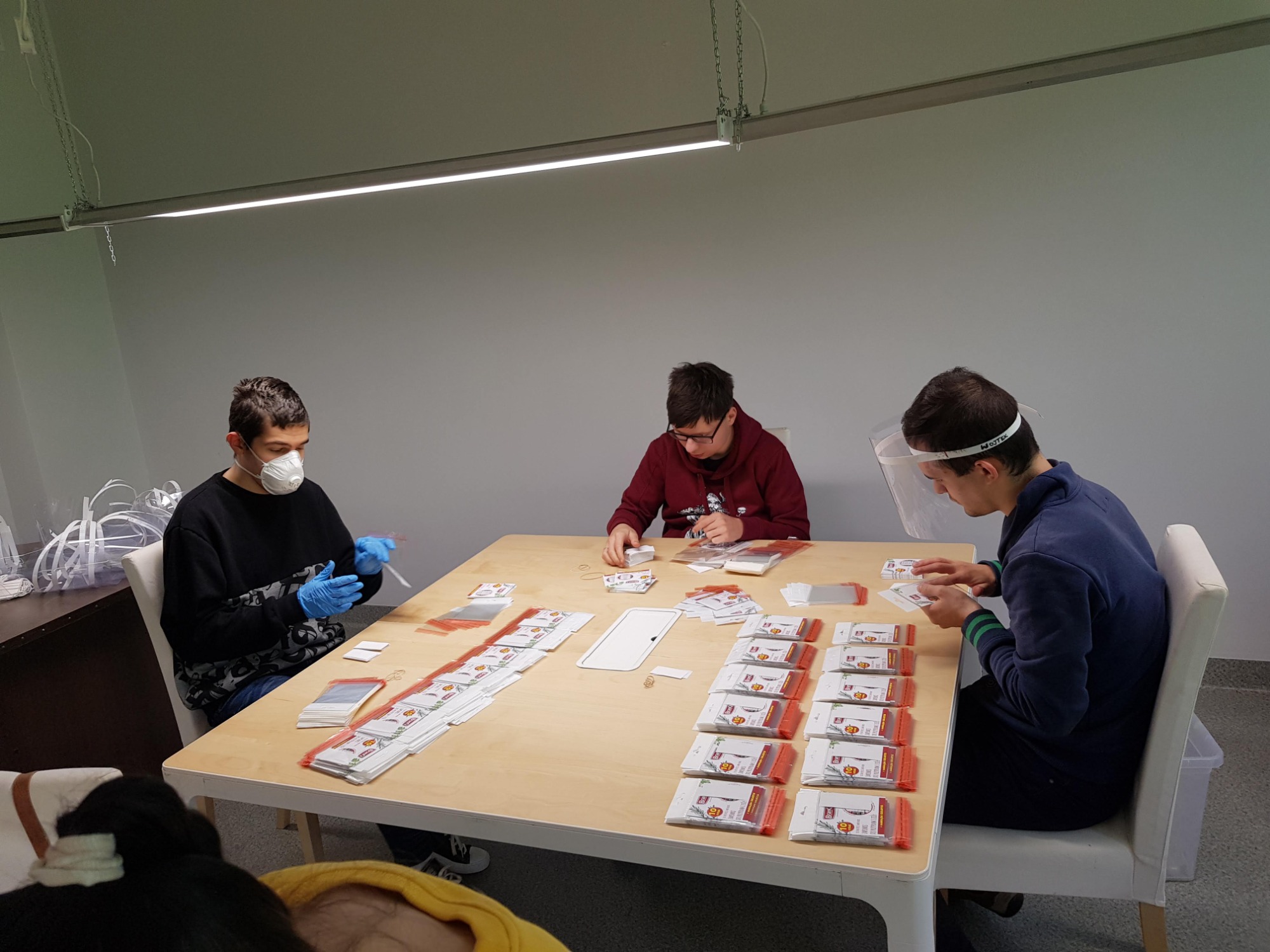 Trzy osoby w maseczkach i rękawiczkach, siedzące przy stole i kompletujące zestawy produktów w przezroczystych opakowaniach, przygotowując je do wysyłki.