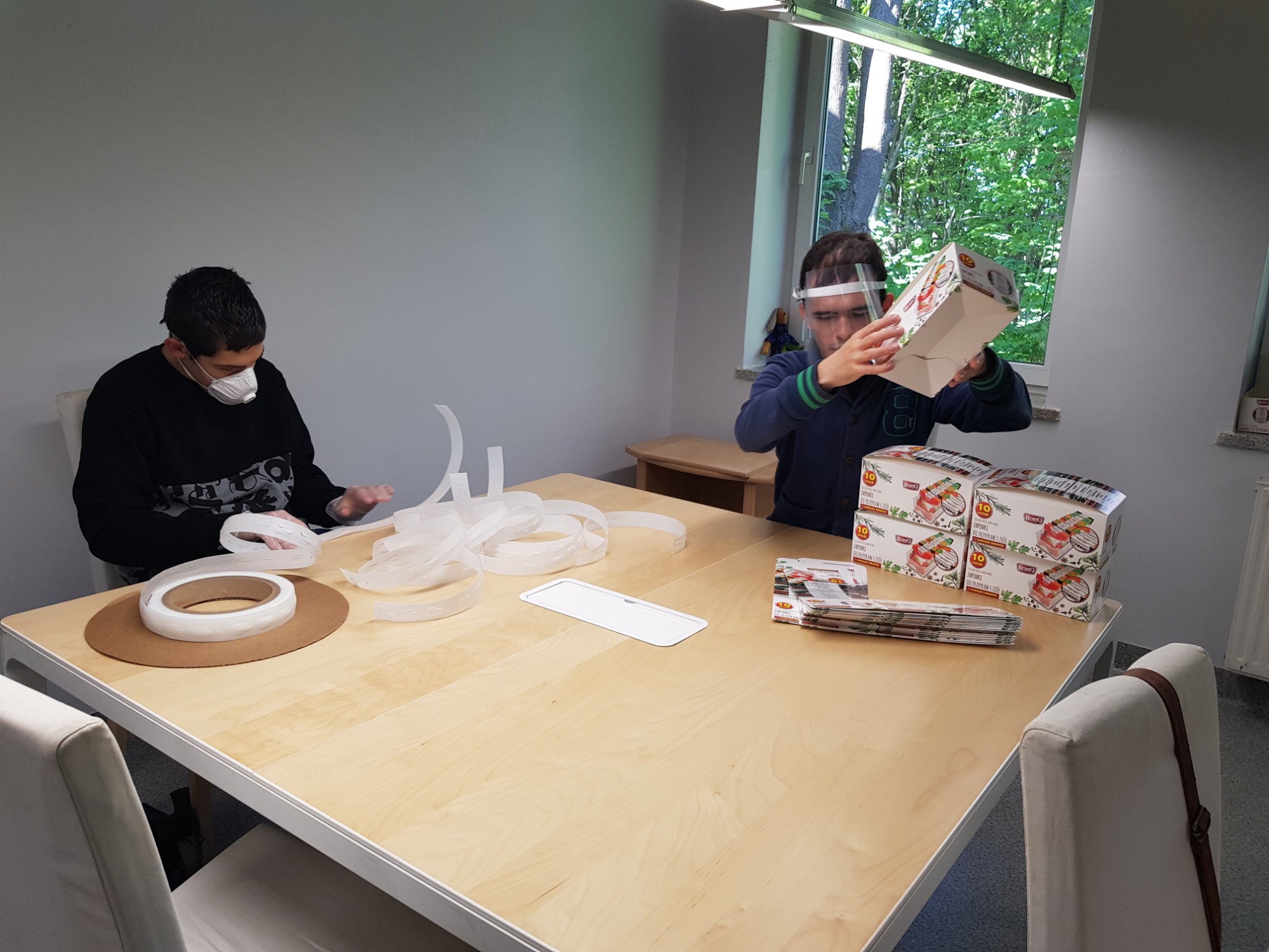 Dwóch mężczyzn w maseczkach i przyłbicy pakuje pudełka z produktami spożywczymi na stole, używając taśmy z dyspenserem i ulotek reklamowych.