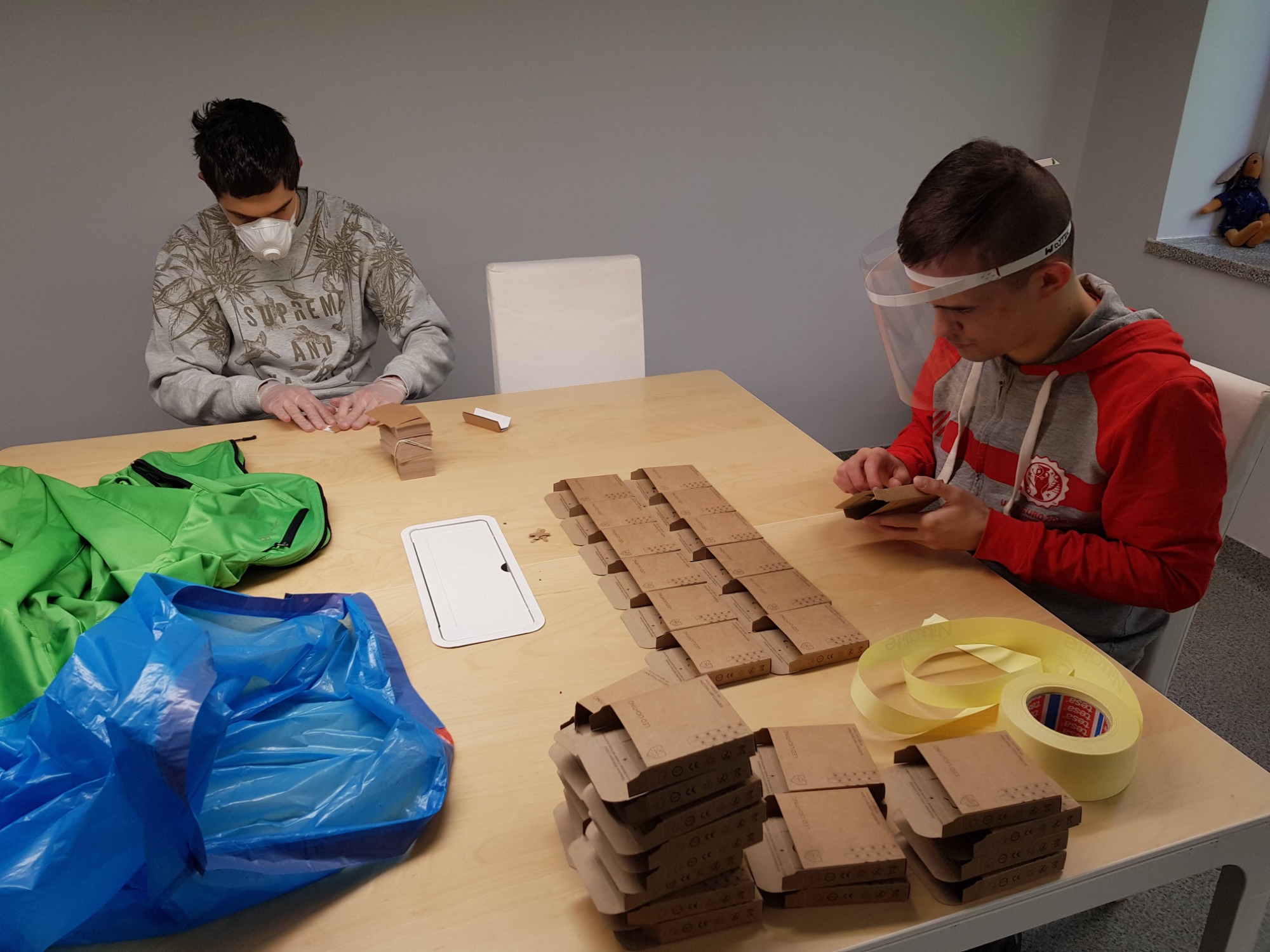 Dwie osoby w maseczkach i przyłbicy składają kartonowe pudełka na stole, obok leżą rolki taśmy klejącej, zapakowane produkty i zielona kurtka.