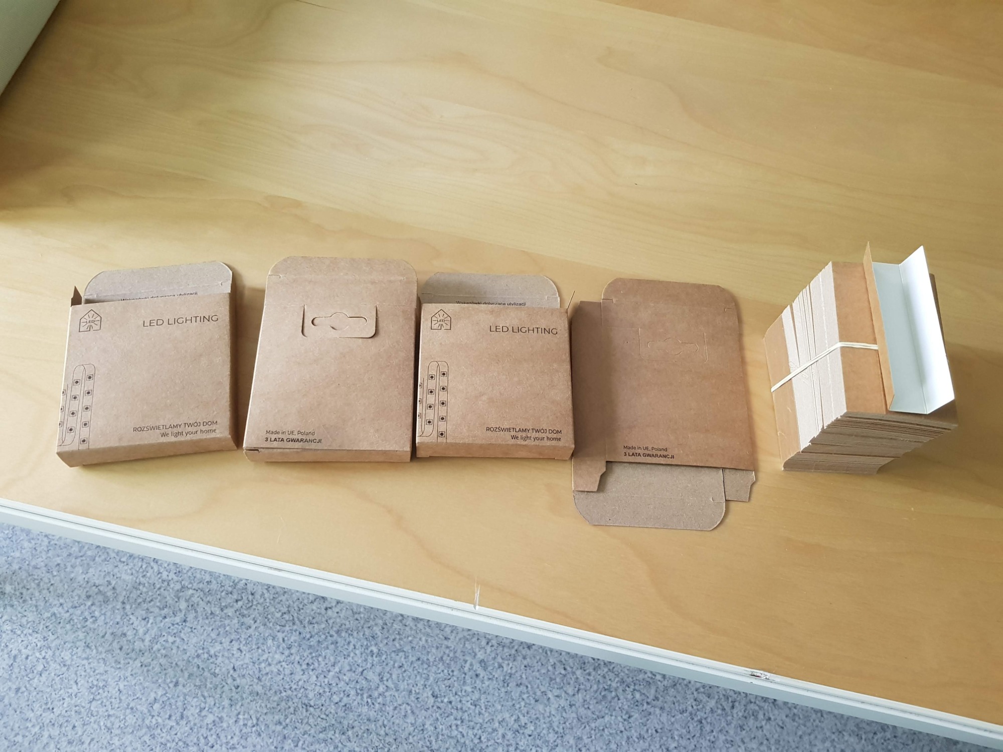 Brązowe, kartonowe opakowania na żarówki LED z nadrukowanym logo i napisem 'Rozświetlamy Twój Dom', ułożone obok stosu pustych, złożonych opakowań związanych gumką.