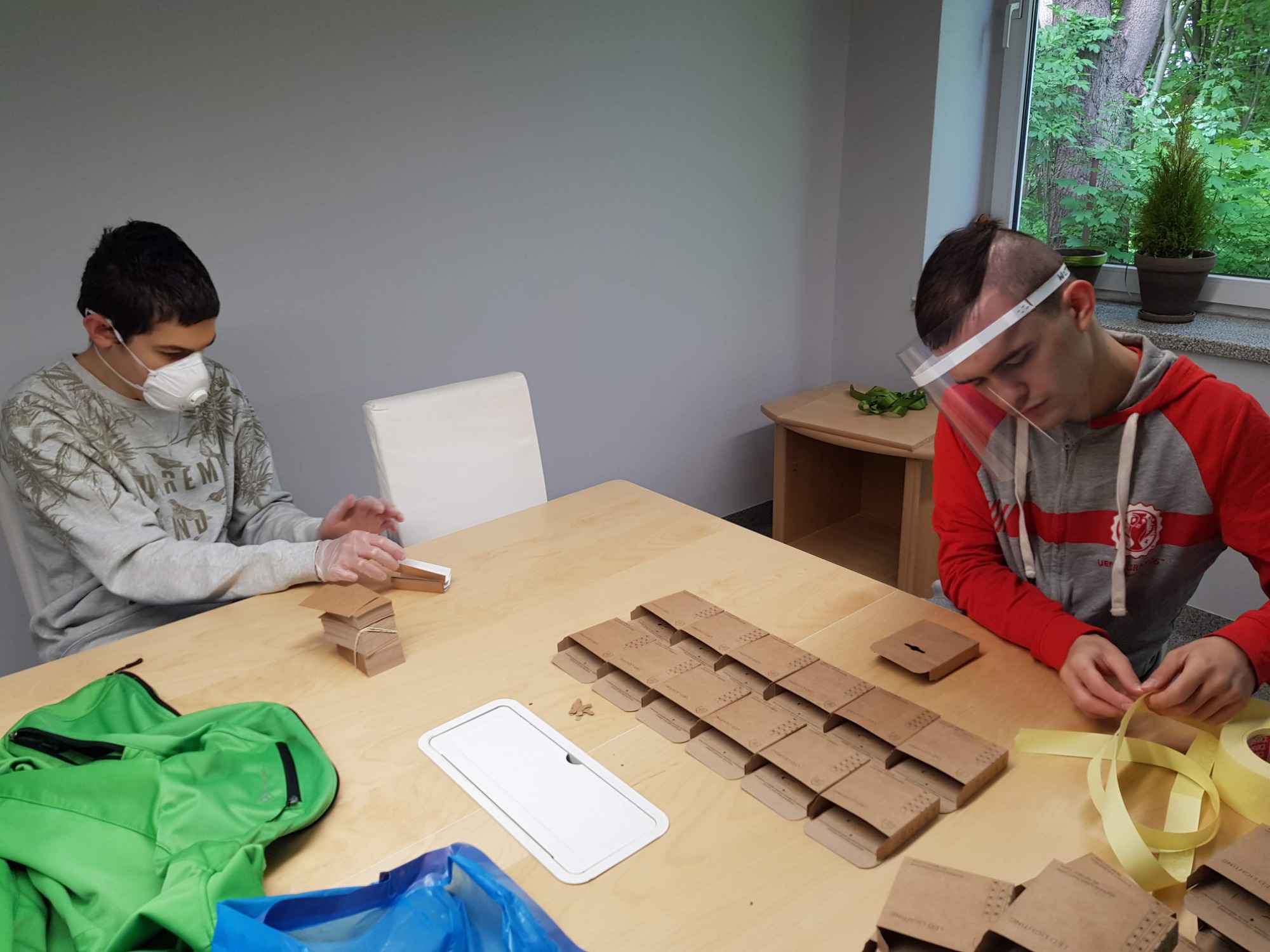 Dwóch młodych mężczyzn w maseczkach i rękawiczkach, przy stole w jasnym pomieszczeniu, składa i przygotowuje kartonowe pudełka z zawieszkami, używając taśmy klejącej.
