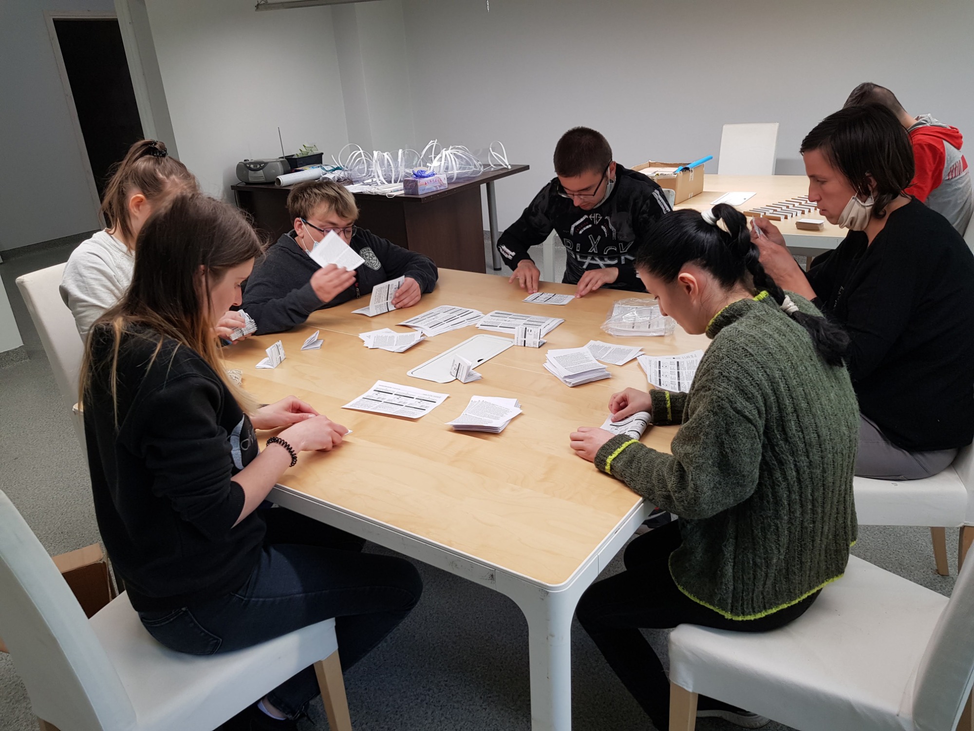 Grupa osób przy stole, składająca ulotki lub instrukcje, prawdopodobnie w ramach procesu kompletowania zestawów. W tle widoczne przezroczyste osłony twarzy.