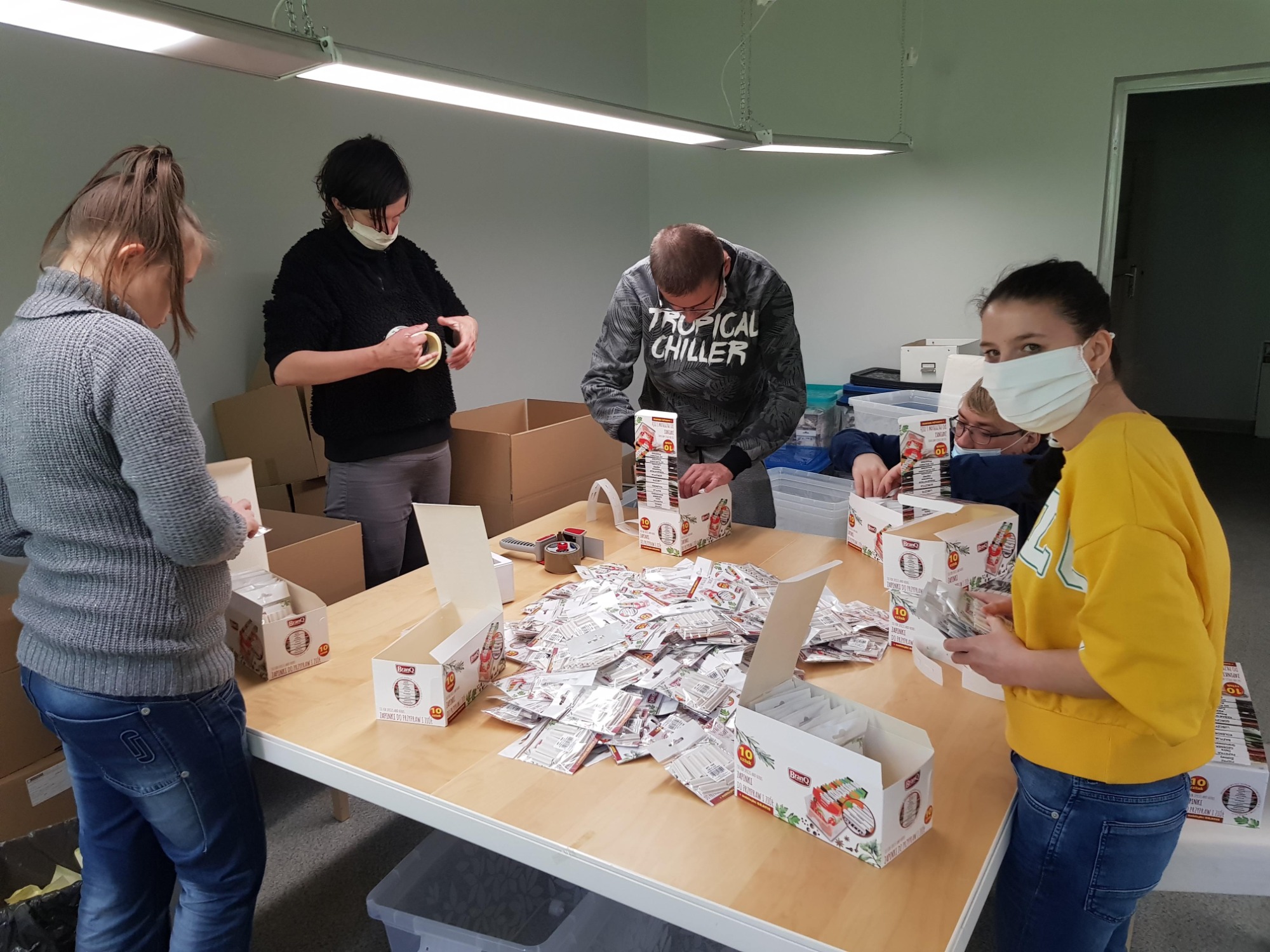 Grupa osób w maskach kompletuje zestawy przypraw na stole, przygotowując je do wysyłki w kartonowych pudełkach.