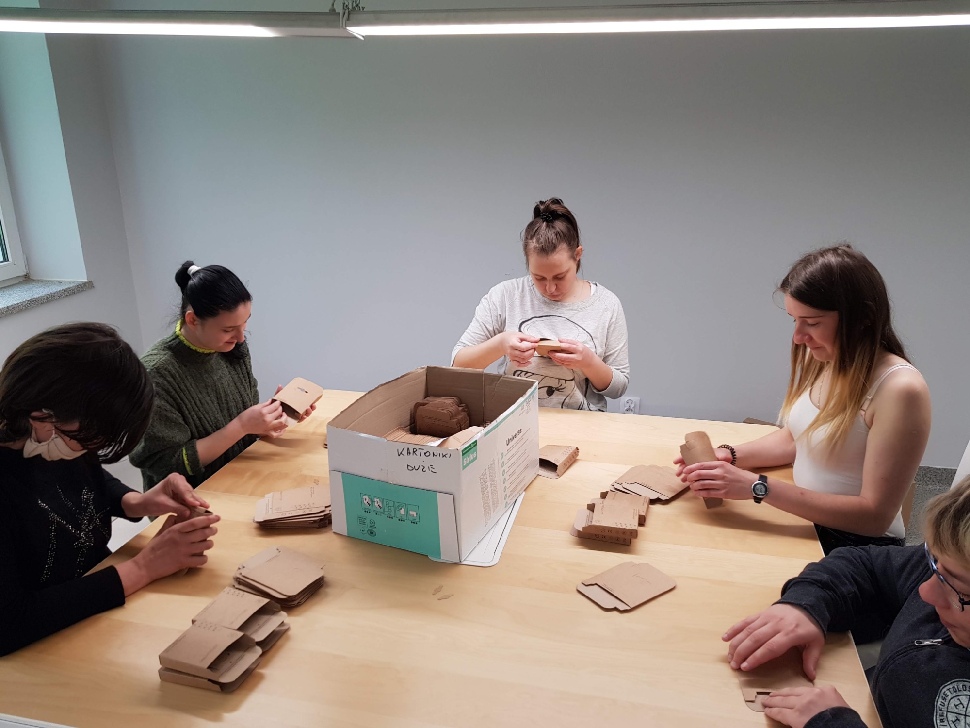 Grupa osób przy stole składa tekturowe pudełka z brązowego papieru, część pudełek znajduje się w kartonie z napisem 'KARTONIKI DUŻE'.