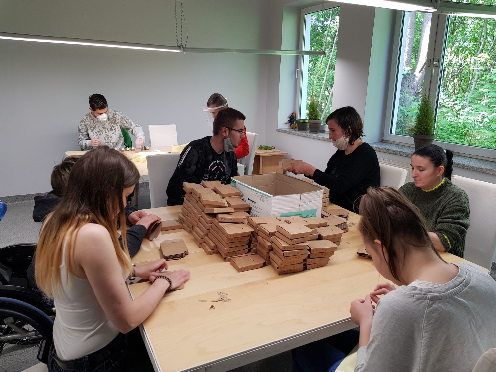 Grupa osób, niektóre w maseczkach, siedzi przy dużym stole składając małe tekturowe pudełka. W tle widoczne okno z zielenią.