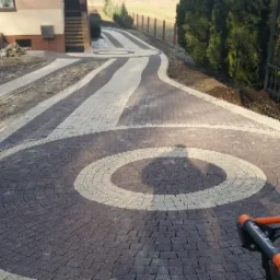 Ułożona dwukolorowa kostka brukowa tworząca okrągły wzór na podjeździe, widoczna brama wjazdowa, w tle budynek i zieleń, częściowo widoczna zagęszczarka wibracyjna.