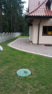 Ułożona ścieżka z kostki brukowej w odcieniach szarości i beżu, obrzeże z jasnej kostki, widok z trawnika na dom z czerwonym dachem i betonowe ogrodzenie w tle, zielona studzienka kanalizacyjna...