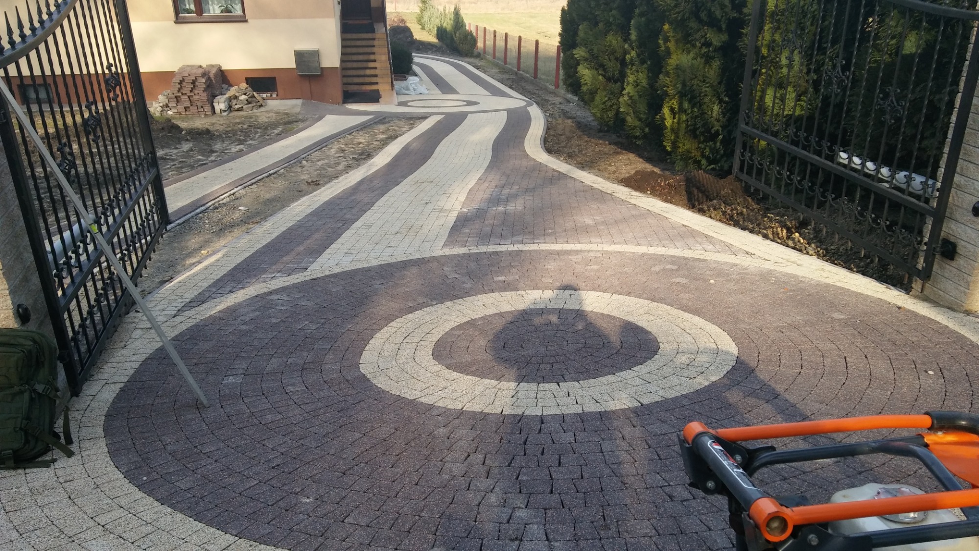 Ułożona dwukolorowa kostka brukowa tworząca okrągły wzór na podjeździe, widoczna brama wjazdowa, w tle budynek i zieleń, częściowo widoczna zagęszczarka wibracyjna.
