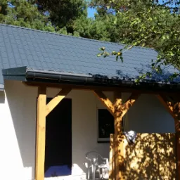 Biały budynek z szarą blachodachówką, drewnianą konstrukcją werandy i czarnymi drzwiami, otoczony zielenią drzew i krzewów.