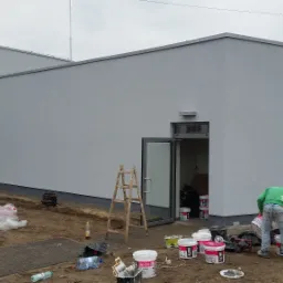 Praca na zewnątrz budynku: Mężczyzna w zielonej bluzie i jeansach przygotowuje farbę przy ścianie budynku pomalowanej na szaro, widoczne puszki z farbą, drabina i otwarte drzwi.