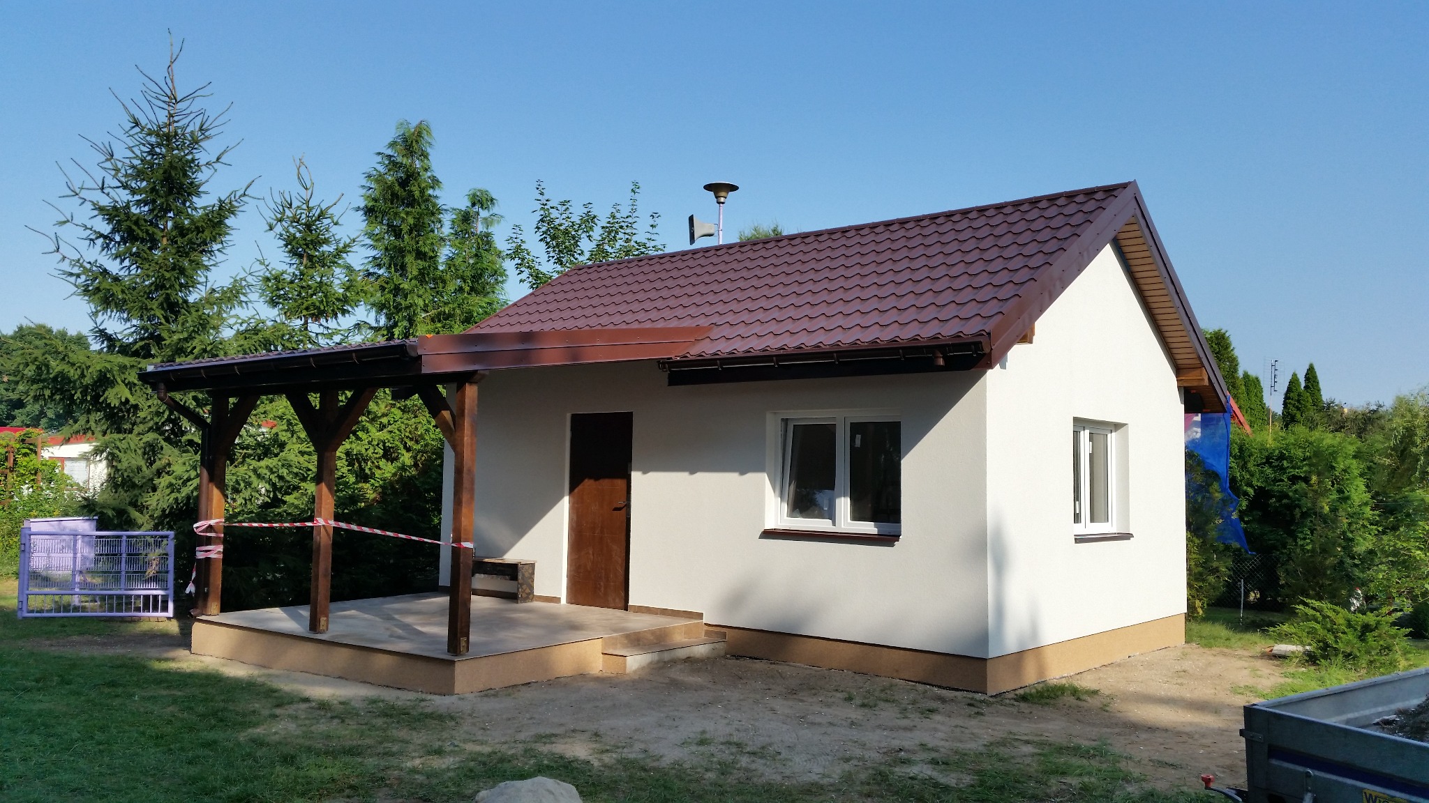 Nowo otynkowany budynek gospodarczy z brązowym dachem i drewnianą werandą, otoczony zielenią i zabezpieczony taśmą ostrzegawczą.