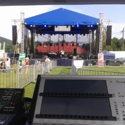Scena z niebieskim zadaszeniem i oświetleniem, widoczna zza konsolety mikserskiej Soundcraft Vi1, w tle ludzie i zielona trawa.
