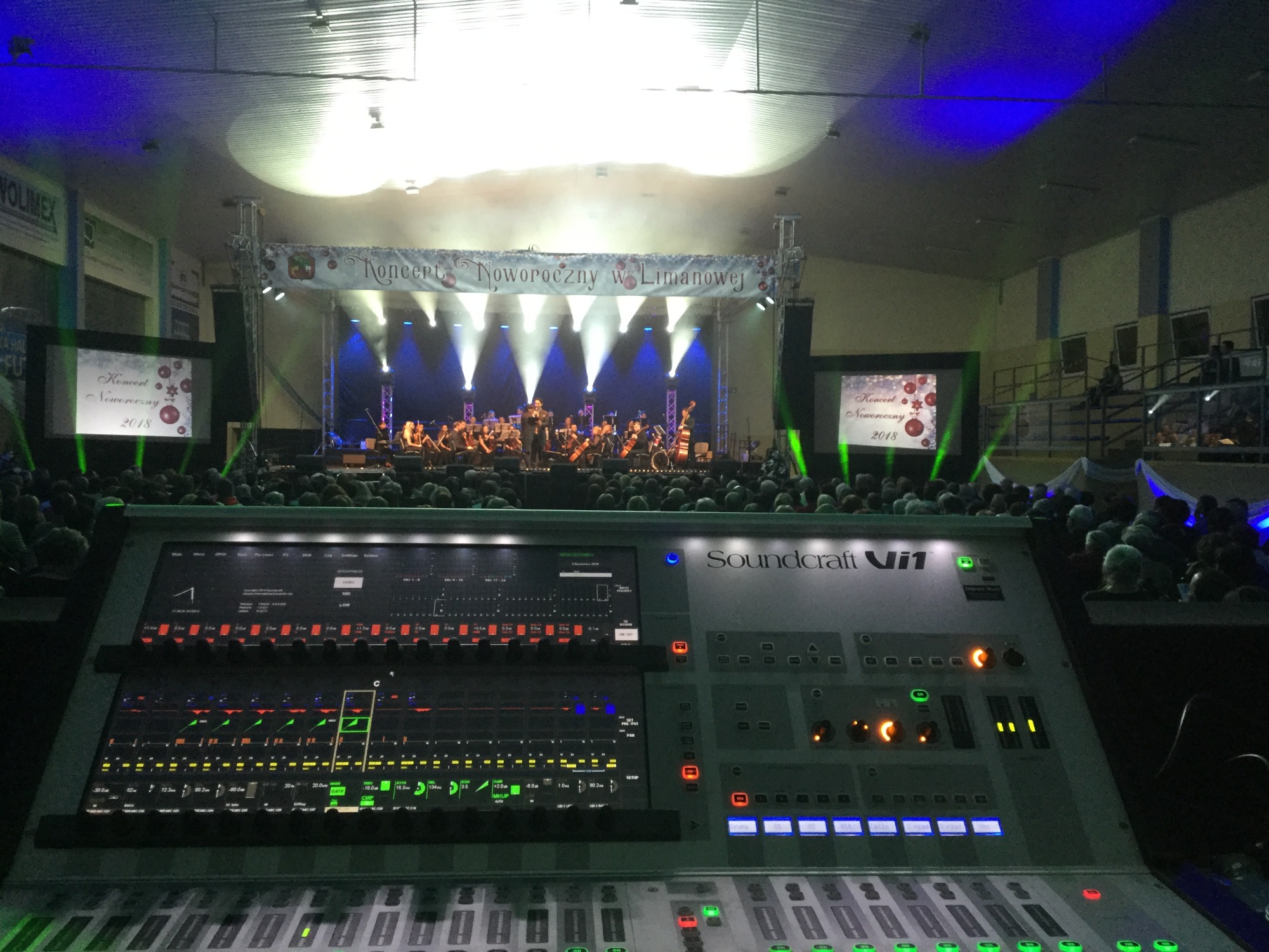 Koncert Noworoczny w hali sportowej, widok zza konsolety mikserskiej Soundcraft Vi1 na oświetloną scenę z orkiestrą i publicznością.